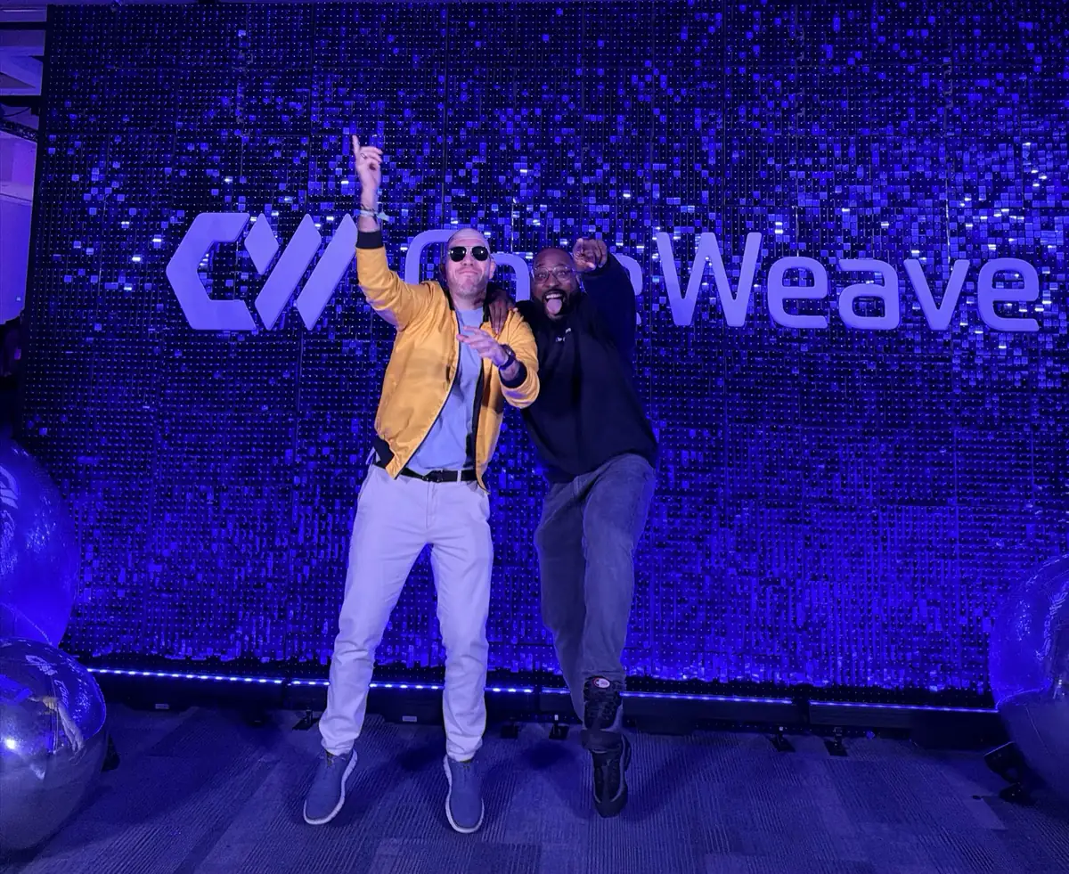 Alex Volkov in a yellow jacket jumping in front of the glittering CoreWeave wall at Fully Connected