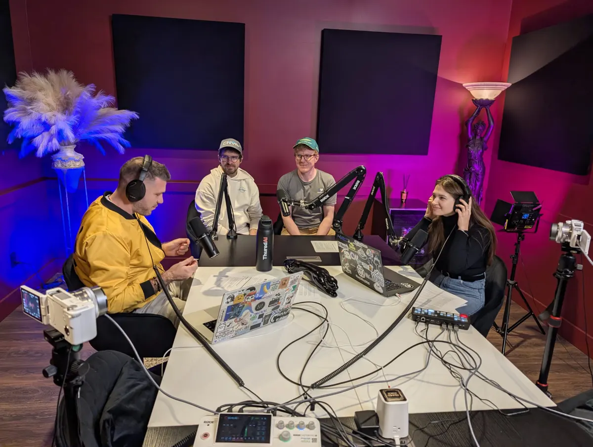 Four people around a table in a podcast studio with red walls, microphones, cameras and laptops